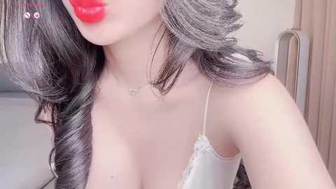 Media: Video of a fair-skinned woman with long, wavy black hair, wearing a white lace-trimmed bralette, red lipstick, and a delicate gold necklace. Background includes a beige wall and a partially visible bed.