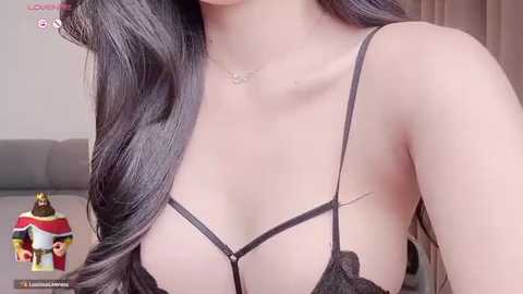 Media: Video of a light-skinned woman with long black hair, wearing a black lace bra, showcasing her cleavage and collarbone. Background includes a gray couch and a window with beige curtains.