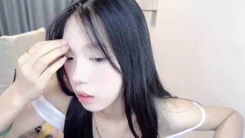 Media: Video of a young Asian woman with long black hair, wearing a white off-shoulder top, touching her forehead with her right hand. She has a pale complexion and looks distressed. Background features a beige sofa and grey curtains.