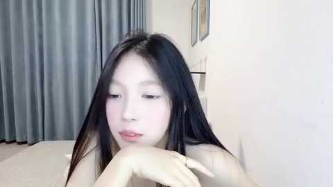 Media: Video of an Asian woman with long black hair, fair skin, and a slim figure, resting her chin on her hands, wearing a light-colored top, in a room with grey curtains and white walls.