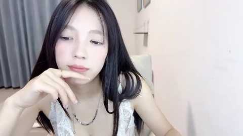 Media: Video of a young East Asian woman with long black hair, wearing a white lace dress, sitting in a minimalist room with white walls and gray curtains.