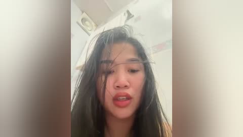 Media: A close-up video of an Asian woman with long, wet black hair, partially covering her face, in a bathroom with white tiles and a ceiling light.