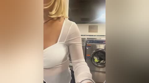 Media: Video of a blonde woman with shoulder-length hair, wearing a white, sheer top, standing beside a modern, stainless steel washing machine in a laundry room.