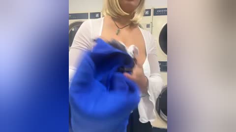 Media: Video of a blonde woman in a white blouse, partially exposing her breast, holding a blue item, in a laundromat with washers and dryers in the background.