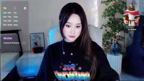 Media: Video of an Asian woman with long black hair, wearing a colorful \"Hello Kitty\" t-shirt, seated in a modern room with blue lighting, a plant, and a gaming setup visible.