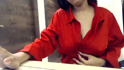 Media: A video of a woman with light skin, wearing a red button-down shirt, partially unbuttoned to reveal cleavage, sitting at a table with a wooden backdrop.