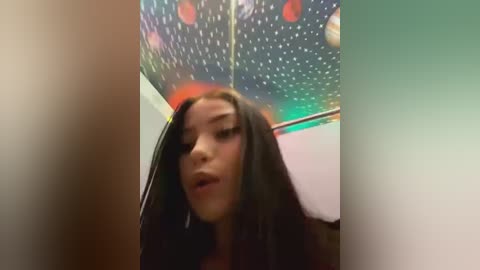 Media: Video of a young woman with long black hair, wearing a black top, standing in a dimly lit room with a colorful, pixelated ceiling.