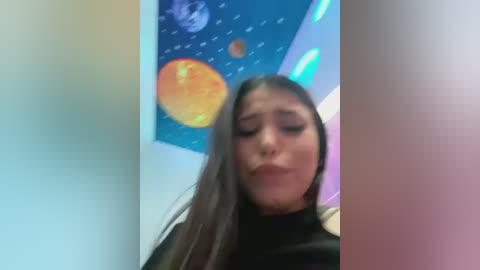 Media: A video of a young woman with long, straight brown hair, wearing a black top, looking down and puckering her lips. Background features a colorful, abstract mural with planets.