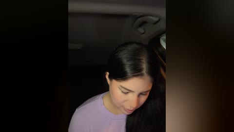 Media: Video of a young woman with long black hair and fair skin, wearing a light purple top, looking down in a dimly lit car interior.