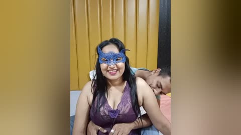 Media: A video of a smiling South Asian woman in a lacy blue mask, wearing a purple lace top, lying on a bed with a man in a blue shirt, behind her.