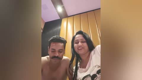 Media: Video of a shirtless South Asian man with a mustache, and a South Asian woman with long black hair, smiling indoors against a yellow and black backdrop.