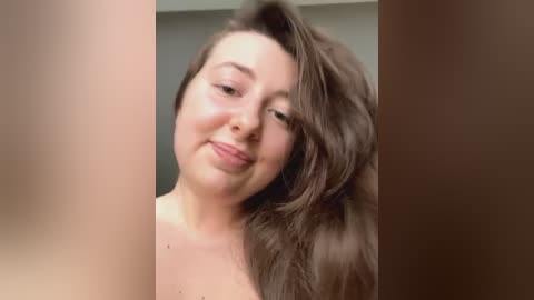 Media: A video of a smiling, fair-skinned woman with long, wavy brown hair, partially obscured by a blurred, brown vertical object. She appears to be indoors, likely in a bathroom.