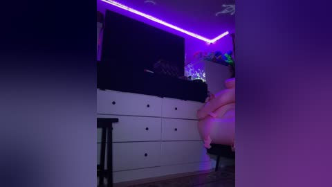 Media: Video of a dimly lit room with purple LED strip lighting. A person with light skin tone sits on a plush, pink chair. White dresser with black handles and a black cushioned headboard in the background.