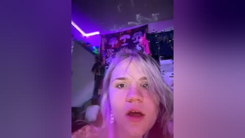 Media: A video of a young woman with platinum blonde hair, wearing a pink top, taken in a dimly lit, messy bedroom with neon purple lights, posters, and a bed in the background.