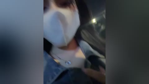 Media: Video of a person wearing a white surgical mask, partially obscured by a car's steering wheel. The person has light skin and is wearing a blue shirt. The background includes dimly lit car interior.