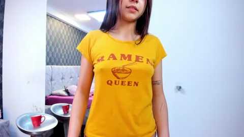 Media: Video of a young woman with straight dark hair, wearing a yellow t-shirt with \"Ramen Queen\" text and a bowl graphic, standing in a modern, white-walled room with a tufted gray couch and red cups on a table.