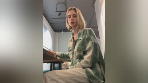 Media: Video of a young woman with shoulder-length blonde hair, wearing a green and white plaid shirt and beige pants, seated in a modern, dimly lit room with white walls and industrial-style ceiling lights.