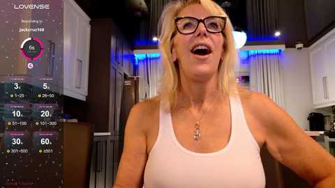 Media: A video of a middle-aged Caucasian woman with blonde hair, wearing glasses, a white tank top, and a necklace, standing in a modern kitchen with blue LED lighting, displaying health stats on a screen.