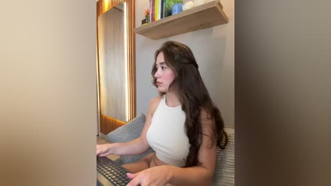 Media: Video of a young woman with long dark hair, wearing a white crop top, sitting at a desk with a keyboard. Background includes a wooden shelf with books and a framed mirror.