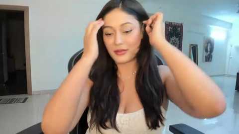 Media: Video of a young, light-skinned Asian woman with long, wavy black hair, wearing a white sleeveless top, adjusting her hair in a modern, brightly lit room with white walls and minimalist decor.