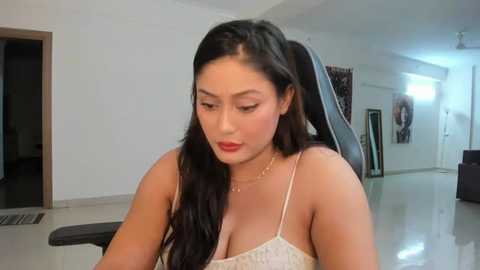 Media: Video of a young Asian woman with long black hair, wearing a white lace camisole, sitting in a modern, minimalist living room.