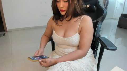 Media: Video of a voluptuous, light-skinned woman with shoulder-length brown hair in a white lace dress, sitting in a black office chair, holding a colorful calculator.