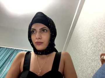 Media: Video of a woman with light skin, wearing a black hijab and sleeveless black top, standing in a room with patterned wallpaper and teal curtains.