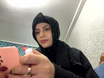 Media: A video of a young woman with light skin, wearing a black hijab and black jacket, holding a pink phone with a white case, in a room with teal and white patterned wallpaper.