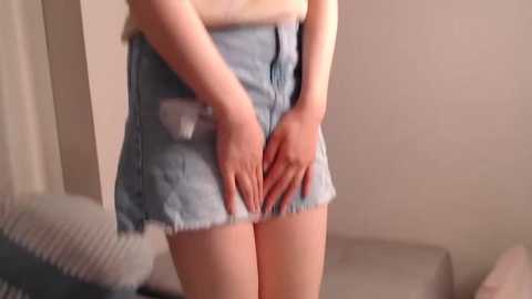 Media: Video of a light-skinned person in light blue denim shorts with frayed edges, hands covering crotch area, standing indoors, beige walls, and a bed with a textured pillow in the background.