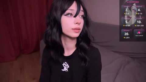 Media: Video of a young woman with long black hair, fair skin, and a slim physique, wearing a black long-sleeve shirt. She stands against a red curtain in a dimly lit room with a gaming monitor displaying a video game interface.