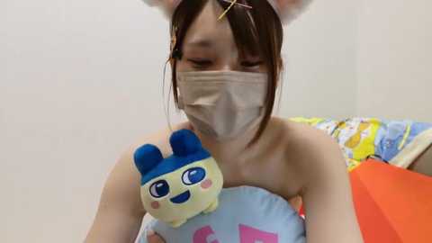 Media: A video of an Asian woman with shoulder-length brown hair, wearing bunny ears and a beige mask, hugging a plush toy with blue ears. She's indoors, surrounded by colorful bedding and toys.