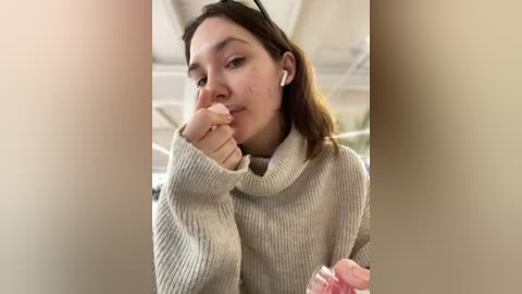 Media: Video of a young woman with light skin, straight brown hair, and glasses, wearing a cream-colored, ribbed knit sweater. She's indoors, possibly in a classroom, with white walls and a window in the background.