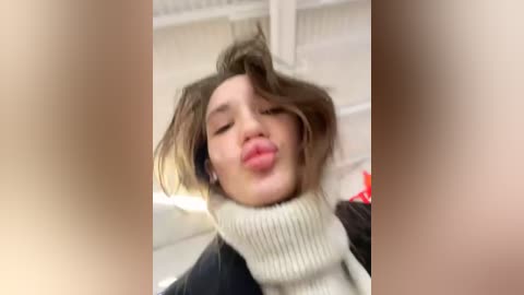 Media: Video of a young woman with tousled brown hair, making a puckered lip kiss expression, wearing a beige turtleneck sweater and black jacket, in a blurry indoor setting.