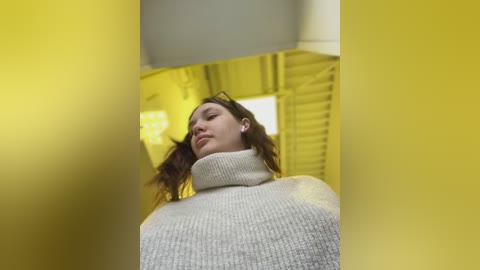 Media: Video of a young woman with long brown hair, wearing a light grey turtleneck sweater, standing in a yellow-lit, modern indoor setting.