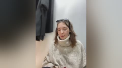 Media: Video of a young woman with light skin and shoulder-length brown hair, wearing a beige turtleneck sweater and black-framed glasses, sitting in a dimly lit, beige-walled changing room.