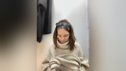 Media: Video of a young woman with shoulder-length brown hair, wearing dark sunglasses and a beige sweater, sitting in a changing room, surrounded by clothes.