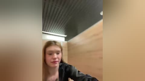 Media: A video of a young woman with light skin and long blonde hair, wearing a black jacket, standing in a modern, light-colored wooden cubicle with a corrugated ceiling.