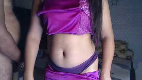 Media: Video of a slender, light-skinned woman with long black hair, wearing a purple satin camisole and matching panties, standing in a dimly lit bedroom with patterned wallpaper.