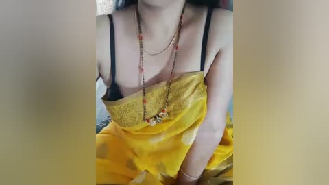 Media: Video of a light-skinned woman with long dark hair, wearing a yellow sari with black lace trim, black bra straps, and a multi-colored beaded necklace. She sits on a bed with a blurred background.