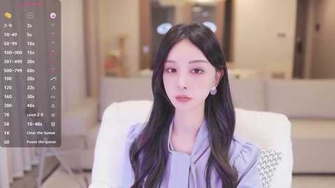 Media: Video of an East Asian woman with long black hair and fair skin, wearing a light blue blouse, sitting on a white couch in a modern, minimalistic living room.