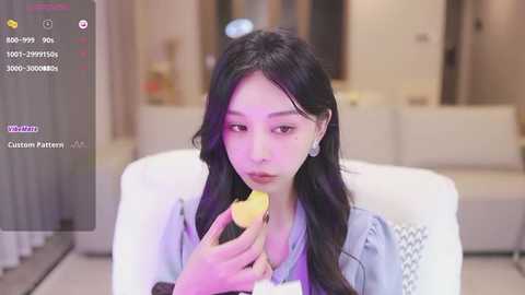 Media: Video of an East Asian woman with long black hair, wearing a light blue shirt, eating a yellow apple in a modern, softly lit living room.