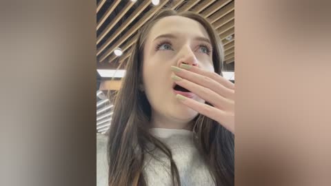 Media: Video of a young woman with long brown hair, light skin, and pale green nail polish, startled, covering her mouth with her hand, in a modern, wooden ceiling setting.