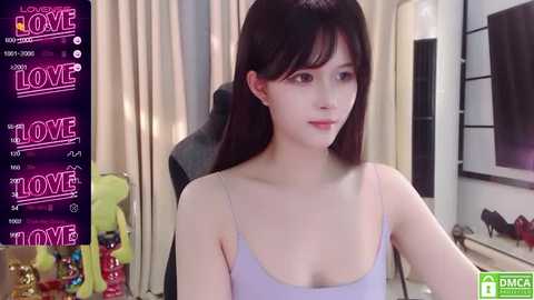 Media: A video of a young Asian woman with fair skin and long dark hair, wearing a light lavender tank top, seated at a desk in a modern room with beige curtains and a black gaming chair.