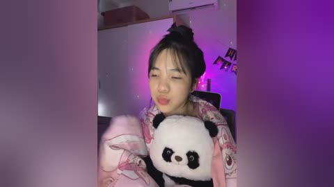 Media: A video of an Asian woman with long black hair tied in a bun, wearing a pink panda-patterned pajama top, hugging a plush panda. The room is dimly lit with purple and pink lights, creating a cozy, intimate atmosphere.