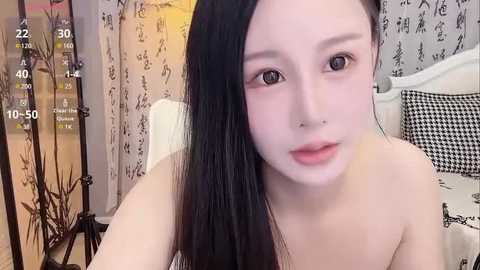 Media: Video of a young, East Asian woman with fair skin, long black hair, and a slender physique. She's topless, sitting on a bed with black and white checkered pillows. The room has Asian-style wallpaper and a wooden screen.