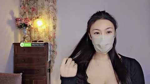 Media: Video of an East Asian woman with long black hair, wearing a face mask, black top, and holding her hair, in a cozy, vintage room with a floral-patterned curtain, wooden dresser, and a glowing lamp.