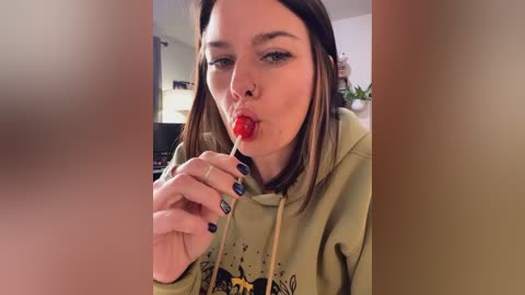 Media: Video of a young woman with light skin and straight brown hair, wearing a green hoodie, licking a red lollipop. Background shows a blurred indoor scene with furniture.