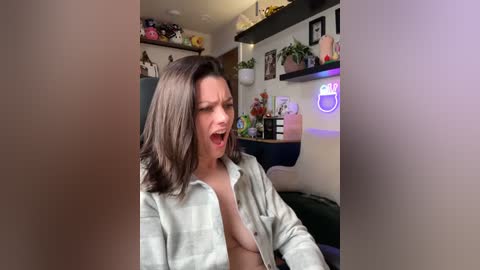 Media: Video of a young woman with light skin and shoulder-length brown hair, wearing a light gray striped shirt open, exposing her small breasts, mid-laugh in a cozy, cluttered room with shelves and plants.