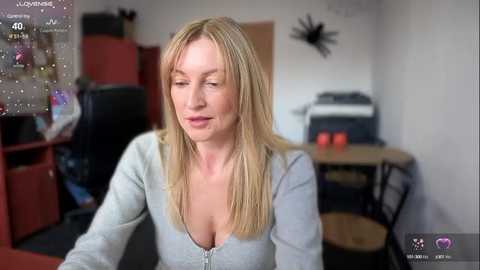 Media: Video of a blonde, middle-aged woman with fair skin, wearing a light gray, low-cut top, sitting in a cluttered office with red shelves, black chair, and a desk.