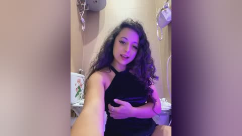 Media: Video of a woman with curly dark hair, wearing a black halter top, in a bathroom with beige tiles, a toilet, and purple lighting.